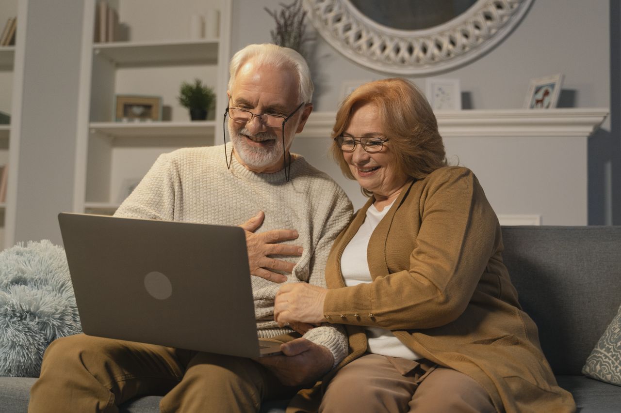 Home Happy senior couple sitting on a sofa having a joyful video call on a laptop in a cozy living room.